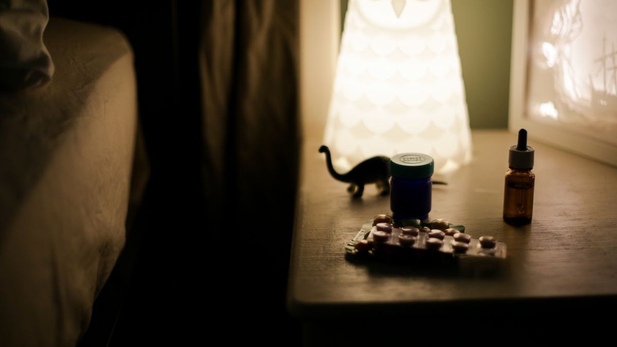 A bedside table at night with a blister pack of tablets, a medicine bottle and a dropper bottle beside a glowing lamp