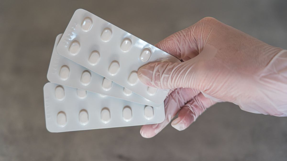 A gloved hand holding three blister strips of white tablets